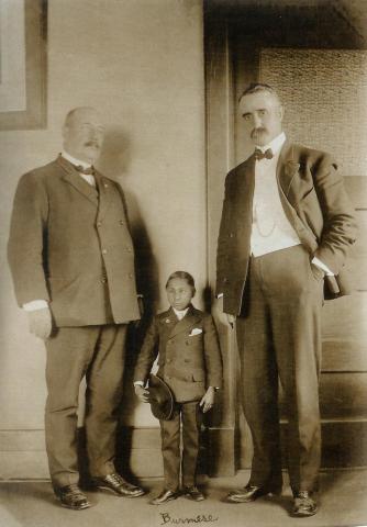The 'Burmese' man stands between two men in suits, the same two men from the 'Russian Giant' picture. The Burmese man stands at waist-height of the men on either side, and he holds a top hat in his hand. He wears a double-breasted suit.